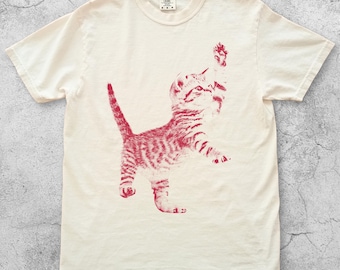 Könnte beinhalten: Elfenbeinfarbenes T-Shirt mit einer roten Illustration eines verspielten Kätzchens. Das Kätzchen ist in einer stehenden Pose mit erhobener Pfote dargestellt. Das Wort "Ivory" ist unten auf dem Shirt aufgedruckt.