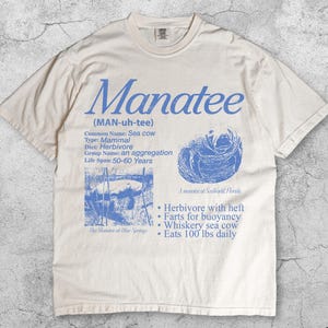 May include: Ivory-colored t-shirt featuring the word "Manatee" in blue, along with informational text and illustrations about manatees. The shirt includes details about their diet, lifespan, and other facts. The text "Ivory" is at the bottom.