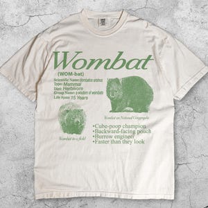 Puede incluir: Camiseta de color marfil con un diseño de wombat verde y texto. El diseño incluye la palabra "Wombat", detalles científicos e ilustraciones del animal. La camiseta también tiene el texto "Wombat in a field" y "Wombat on National Geographic".
