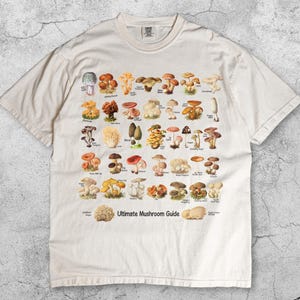 May include: Ivory-colored t-shirt featuring a detailed illustration of various mushrooms. The shirt includes the text "Ultimate Mushroom Guide" and the color "Ivory" at the bottom. The design showcases a variety of mushroom species in vibrant colors.