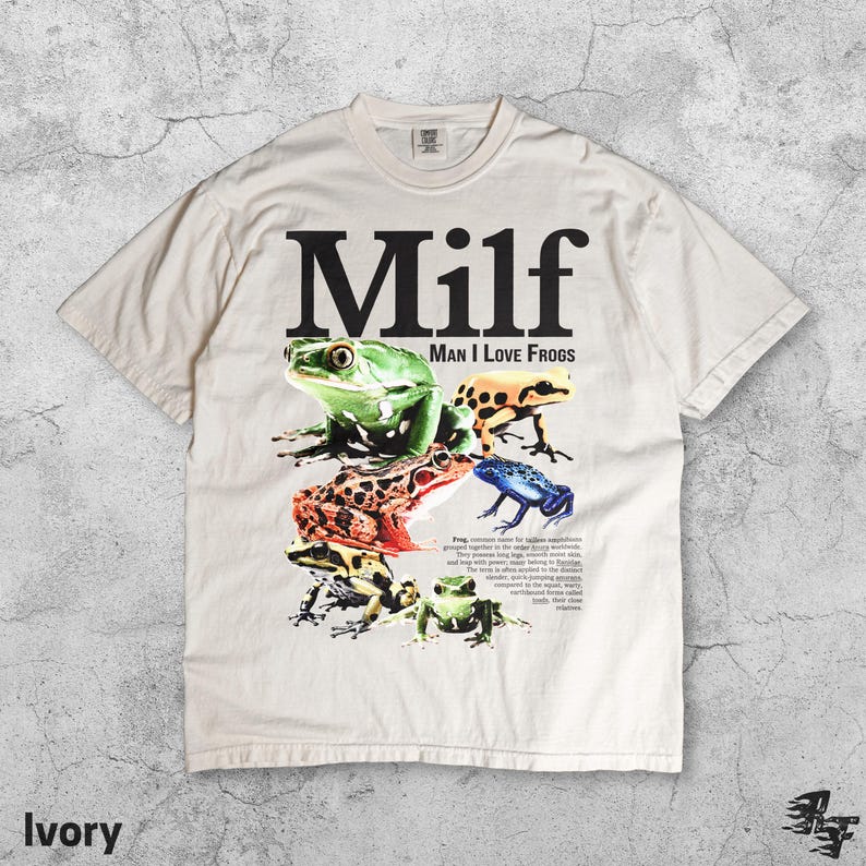May include: Ivory-colored t-shirt featuring the word "Milf" in large black letters above the phrase "Man I Love Frogs." The shirt displays a colorful graphic of various frog species. The word "Ivory" is printed at the bottom.