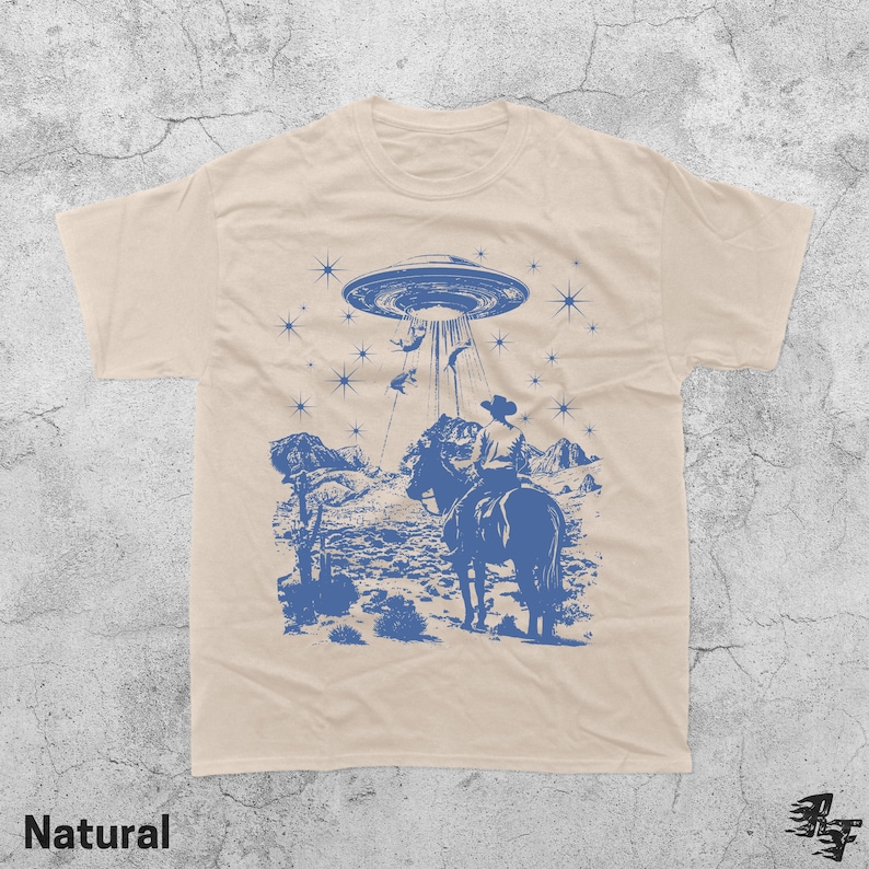 May include: A natural-colored t-shirt featuring a blue graphic of a cowboy on horseback being abducted by a UFO. The design includes a desert landscape, stars, and the word "Natural" at the bottom.
