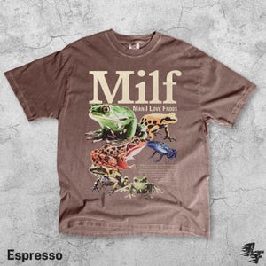 May include: Espresso-colored t-shirt featuring the word "Milf" in large, white letters above a graphic of several colorful frogs. The phrase "Man I Love Frogs" is printed below the word "Milf".