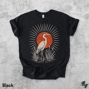 Pu&ograve; includere: Una t-shirt nera a maniche corte con una stampa grafica di un airone bianco in piedi nell'acqua tra i canneti. Dietro l'uccello c'&egrave; un grande sole arancione testurizzato con linee bianche radianti in un motivo circolare.