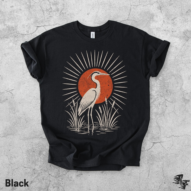 Pu&ograve; includere: Una t-shirt nera a maniche corte con una stampa grafica di un airone bianco in piedi nell'acqua tra i canneti. Dietro l'uccello c'&egrave; un grande sole arancione testurizzato con linee bianche radianti in un motivo circolare.