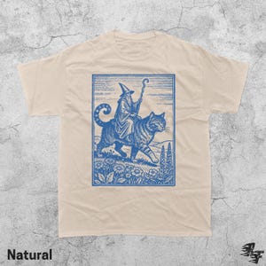 May include: A natural-colored t-shirt featuring a blue illustration of a wizard riding a large cat through a field of flowers. The wizard wears a tall hat and holds a staff. The word "Natural" is printed at the bottom.