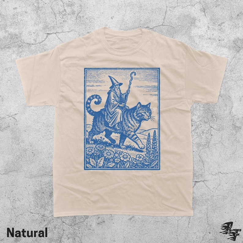 May include: A natural-colored t-shirt featuring a blue illustration of a wizard riding a large cat through a field of flowers. The wizard wears a tall hat and holds a staff. The word "Natural" is printed at the bottom.