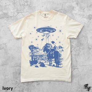 May include: Ivory-colored t-shirt with a blue graphic of a cowboy on horseback being abducted by a UFO. The design includes stars, mountains, and desert plants. The word "Ivory" is printed at the bottom.