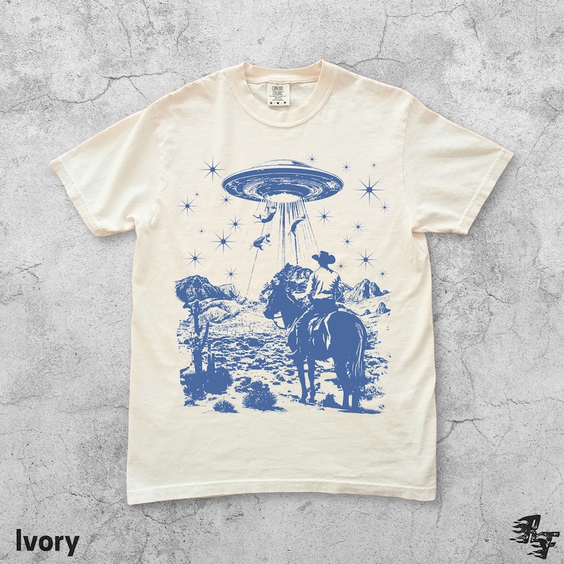 May include: Ivory-colored t-shirt with a blue graphic of a cowboy on horseback being abducted by a UFO. The design includes stars, mountains, and desert plants. The word "Ivory" is printed at the bottom.