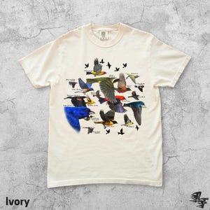 May include: Ivory-colored t-shirt featuring a colorful illustration of various bird species in flight. The design includes detailed depictions of different birds with their names. The shirt is made of a soft material.