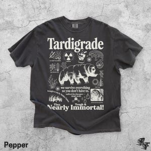 Puede incluir: Una camiseta gris carb&oacute;n de manga corta con un gran gr&aacute;fico blanco de un tard&iacute;grado rodeado de iconos cient&iacute;ficos y textos que incluyen "Tardigrade", "we survive everything so you don't have to" y "Tiny, Nearly Immortal!"