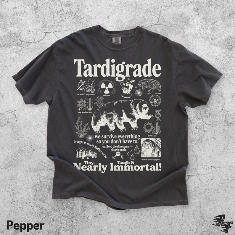 Puede incluir: Una camiseta gris carb&oacute;n de manga corta con un gran gr&aacute;fico blanco de un tard&iacute;grado rodeado de iconos cient&iacute;ficos y textos que incluyen "Tardigrade", "we survive everything so you don't have to" y "Tiny, Nearly Immortal!"