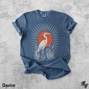 Pu&ograve; includere: Una t-shirt blu a maniche corte con una stampa grafica di un airone bianco in piedi nell'acqua tra i canneti. Dietro l'uccello c'&egrave; un grande sole arancione con linee bianche radianti. La maglietta presenta un girocollo e maniche risvoltate.