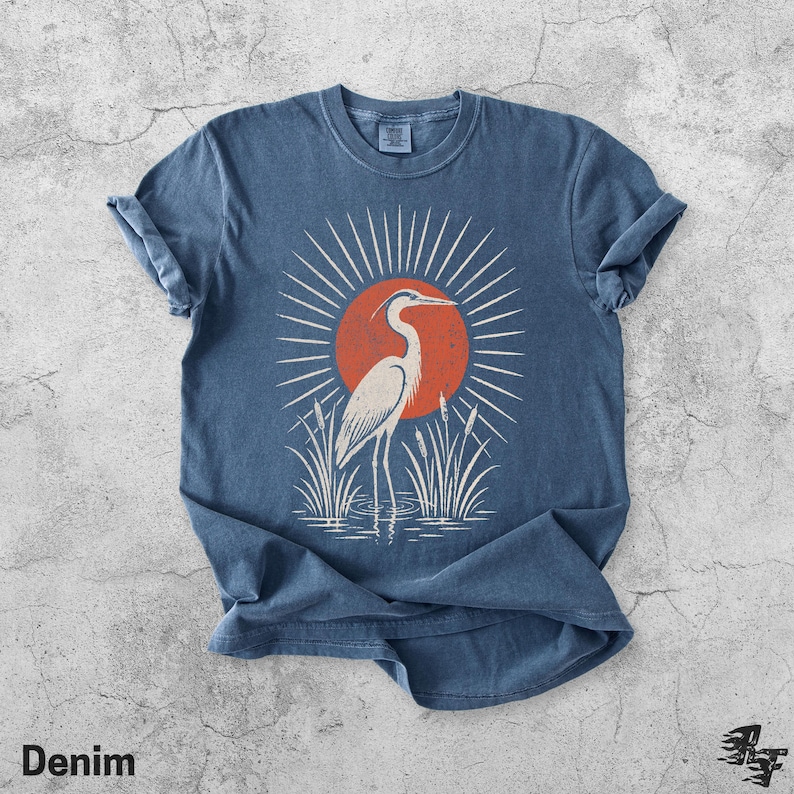 Pu&ograve; includere: Una t-shirt blu a maniche corte con una stampa grafica di un airone bianco in piedi nell'acqua tra i canneti. Dietro l'uccello c'&egrave; un grande sole arancione con linee bianche radianti. La maglietta presenta un girocollo e maniche risvoltate.