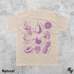 Pu&ograve; includere: T-shirt beige con illustrazione di frutta viola. Il design include uva, una pera, un'arancia a fette, una banana, una mela, una prugna, un melograno e un ananas. La parola "Natural" &egrave; stampata in basso.