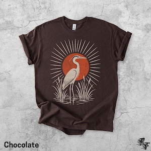 Pu&ograve; includere: Una t-shirt a maniche corte color marrone cioccolato con una stampa grafica di un airone in piedi tra i canneti. Il design include un grande sole arancione con linee radianti dietro l'uccello. La maglietta ha un girocollo.