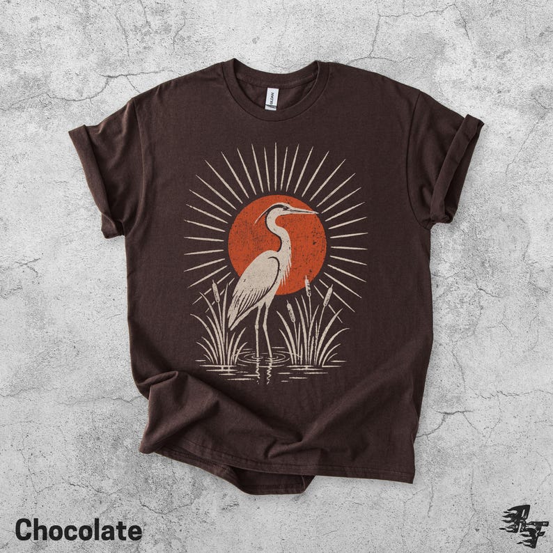 Pu&ograve; includere: Una t-shirt a maniche corte color marrone cioccolato con una stampa grafica di un airone in piedi tra i canneti. Il design include un grande sole arancione con linee radianti dietro l'uccello. La maglietta ha un girocollo.