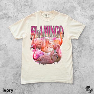 May include: Ivory-colored t-shirt featuring a graphic of several pink flamingos with the word "FLAMINGO" in pink and gold lettering. The shirt is a crew neck and has short sleeves.