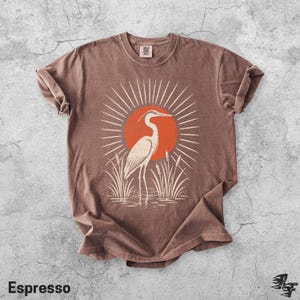 Pu&ograve; includere: Una t-shirt marrone a maniche corte con una stampa grafica di un airone bianco in piedi nell'erba alta davanti a un grande sole arancione con linee bianche radianti. La maglietta ha un girocollo e un'etichetta visibile all'interno.