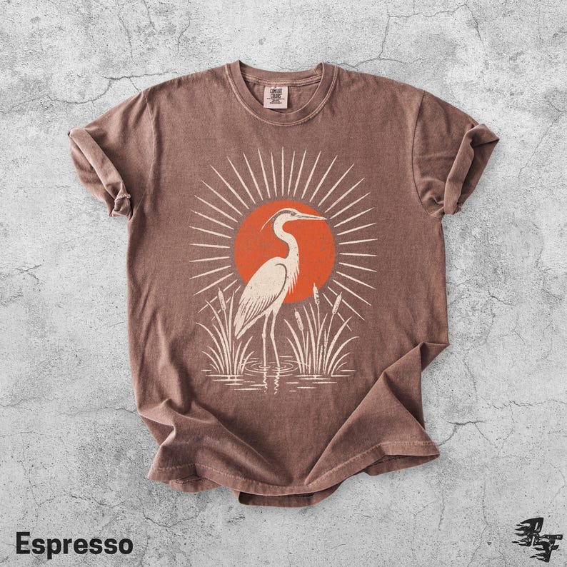 Pu&ograve; includere: Una t-shirt marrone a maniche corte con una stampa grafica di un airone bianco in piedi nell'erba alta davanti a un grande sole arancione con linee bianche radianti. La maglietta ha un girocollo e un'etichetta visibile all'interno.