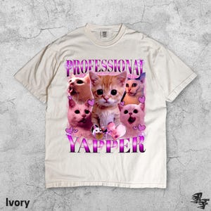 May include: Ivory-colored t-shirt featuring a graphic of several cats with the text "PROFESSIONAL YAPPER" in a purple and white font. The cats have various expressions, and pink hearts are scattered around the design.