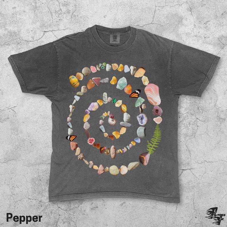 Puede incluir: Camiseta gris oscuro con un dise&ntilde;o en espiral hecho de piedras, conchas y mariposas de colores. La palabra "Pepper" est&aacute; impresa en la parte inferior izquierda.