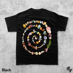 Puede incluir: Camiseta negra con un dise&ntilde;o en espiral hecho de varias piedras de colores, conchas y un helecho. La palabra "Black" est&aacute; impresa en la esquina inferior izquierda de la camiseta.