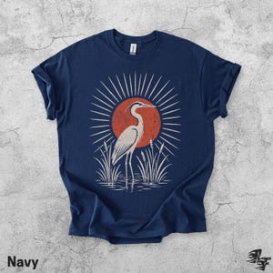 Pu&ograve; includere: Una t-shirt blu navy a maniche corte con una stampa grafica di un airone bianco in piedi nell'acqua tra i canneti. Dietro l'uccello c'&egrave; un grande cerchio arancione pieno con linee bianche a raggiera che si irradiano verso l'esterno.