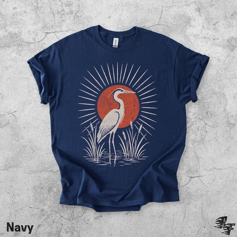 Pu&ograve; includere: Una t-shirt blu navy a maniche corte con una stampa grafica di un airone bianco in piedi nell'acqua tra i canneti. Dietro l'uccello c'&egrave; un grande cerchio arancione pieno con linee bianche a raggiera che si irradiano verso l'esterno.