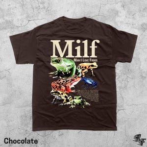 May include: Chocolate-colored t-shirt featuring the word "Milf" in large, white letters above a graphic of several colorful frogs. The text "Man I Love Frogs" is below the word "Milf". The word "Chocolate" is at the bottom.
