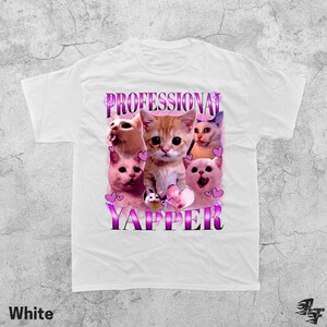 May include: White t-shirt featuring a graphic of several cats with pink hearts and the text "PROFESSIONAL YAPPER" in a purple and pink font. The shirt is made of a soft material.