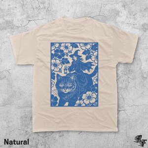 Op de afbeelding: Een natuurlijk gekleurd t-shirt met een blauwe afbeelding van een heks die op een kat rijdt door een bloemenontwerp. De heks draagt een hoed en cape, met een lantaarn. Het woord "Natural" staat onderaan.
