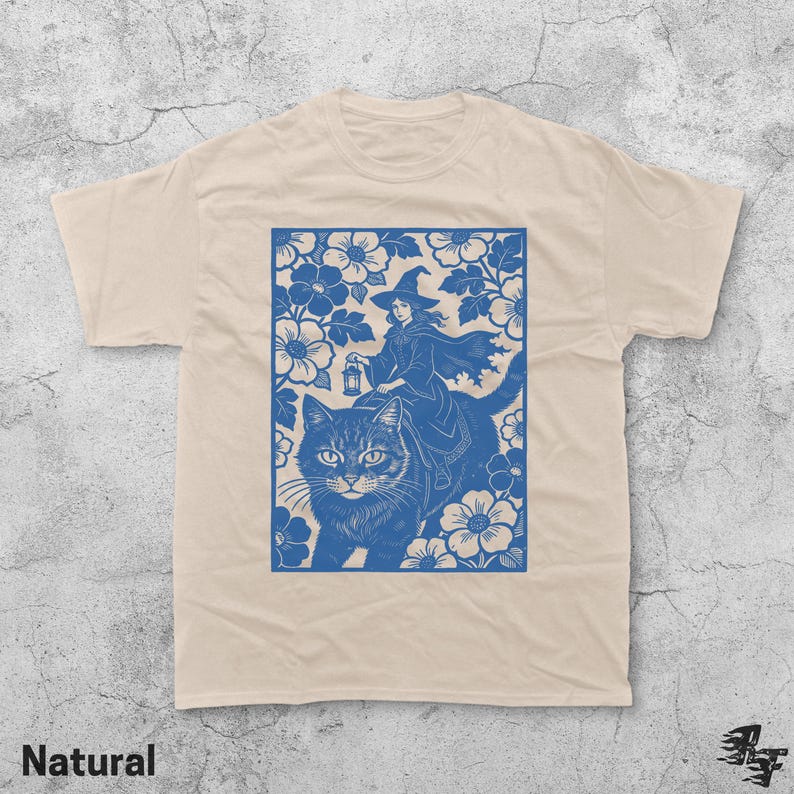 Op de afbeelding: Een natuurlijk gekleurd t-shirt met een blauwe afbeelding van een heks die op een kat rijdt door een bloemenontwerp. De heks draagt een hoed en cape, met een lantaarn. Het woord "Natural" staat onderaan.