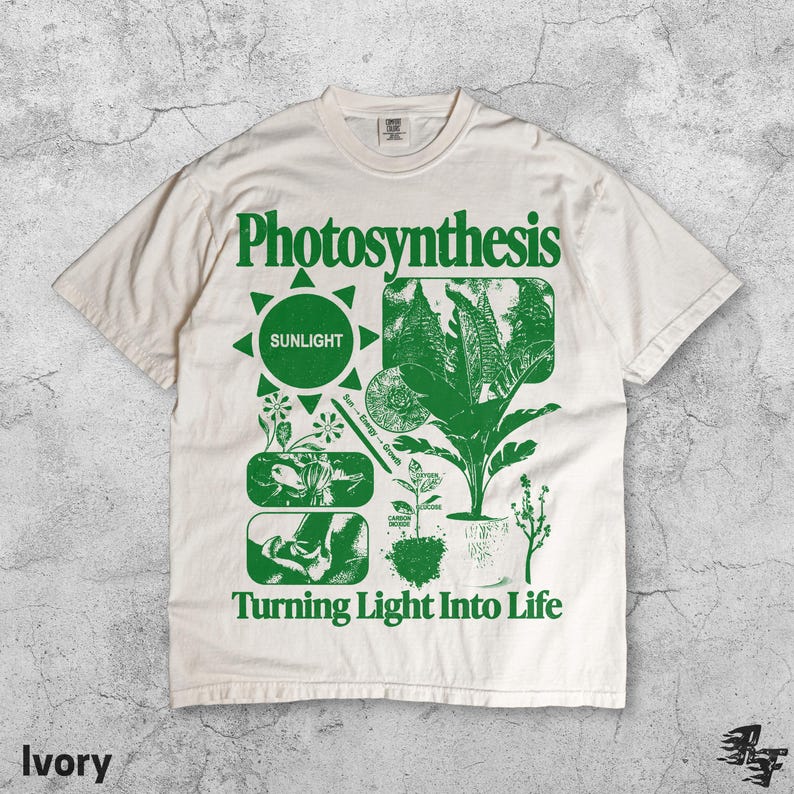 May include: Ivory-colored t-shirt with a green graphic design. The design includes the word "Photosynthesis" above a sun symbol and various plant illustrations. The text "Turning Light Into Life" is printed below the illustrations.
