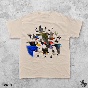 May include: Ivory-colored t-shirt featuring a colorful illustration of various bird species, each labeled with their name. The design includes birds in flight and perched, with a variety of colors and patterns. The word "Ivory" is printed at the bottom.