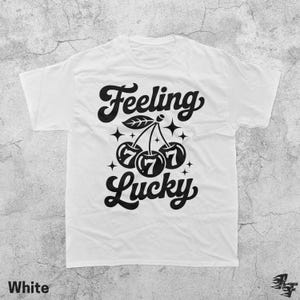 May include: White t-shirt with a black graphic that reads "Feeling Lucky." The design features cherries with the number 7 on each, along with stars and leaves. The word "White" is printed at the bottom.