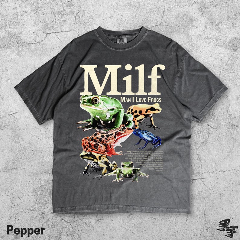 May include: Dark gray t-shirt with the word "Milf" in large white letters above a collection of colorful frog illustrations. The text "Man I Love Frogs" is below the word "Milf". The shirt is made of a soft material.