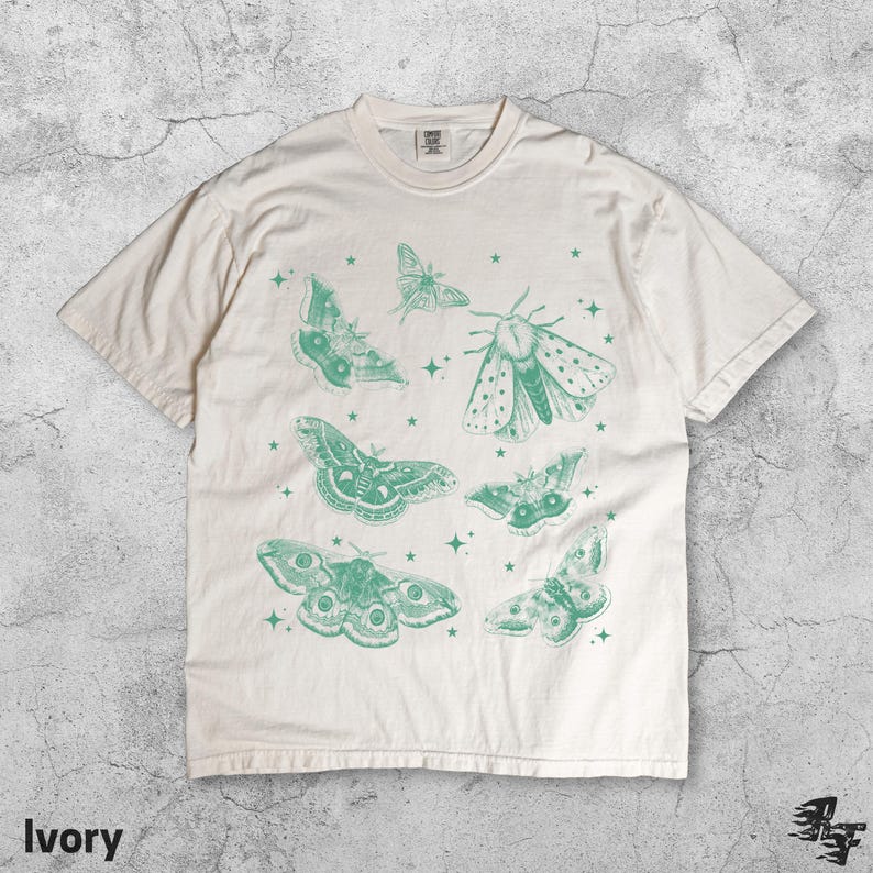 May include: Ivory-colored t-shirt featuring a collection of green moth illustrations and stars. The moths vary in size and design, with intricate wing patterns. The shirt has a classic crew neck and short sleeves, made of a soft, comfortable material.