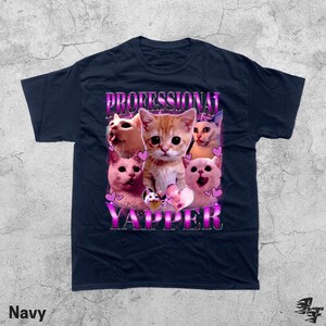 May include: Navy blue t-shirt featuring a graphic of several cats with the text "PROFESSIONAL YAPPER" in a pink and purple font. The cats have various expressions and are surrounded by pink hearts.