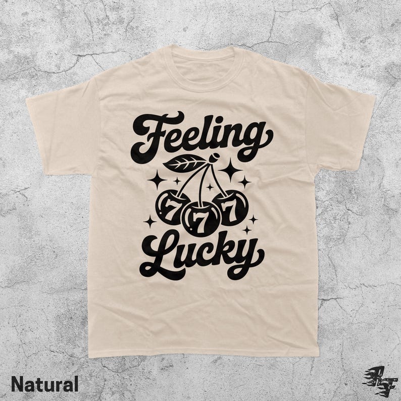 May include: A natural-colored t-shirt with a black graphic design. The design features the words "Feeling Lucky" with cherries and the number 777. The shirt is made of a soft material.