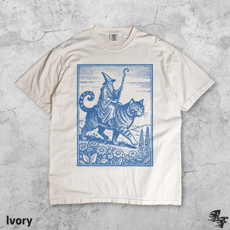 May include: Ivory-colored t-shirt featuring a blue illustration of a wizard riding a large cat through a field of flowers. The wizard holds a staff. The word "Ivory" is printed at the bottom of the shirt.