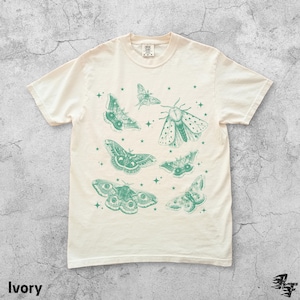 May include: Ivory-colored t-shirt featuring a collection of detailed green moth illustrations and small star accents. The shirt has a classic crew neck and short sleeves. The word "Ivory" is printed at the bottom left.