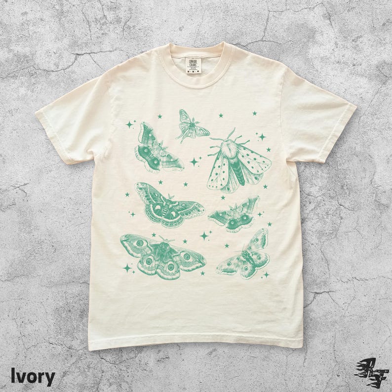 May include: Ivory-colored t-shirt featuring a collection of detailed green moth illustrations and small star accents. The shirt has a classic crew neck and short sleeves. The word "Ivory" is printed at the bottom left.