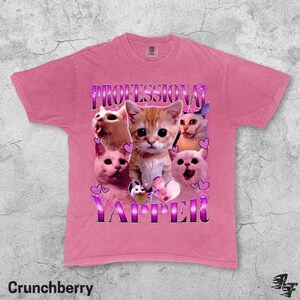 May include: A pink t-shirt with a graphic featuring several cat faces and the words "PROFESSIONAL YAPPER" in a purple, stylized font. The shirt has a crew neck and short sleeves. The word "Crunchberry" is printed at the bottom.