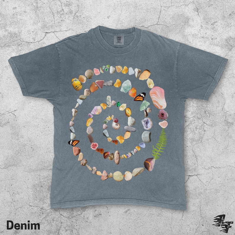 Puede incluir: Camiseta azul denim con un dise&ntilde;o en espiral hecho de piedras, conchas y mariposas. La camiseta tiene cuello redondo y mangas cortas. La palabra "Denim" est&aacute; impresa en la esquina inferior izquierda.