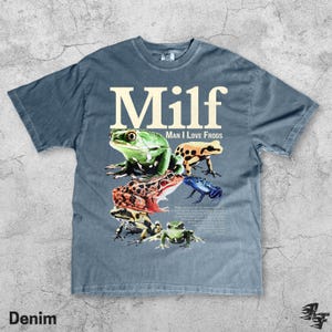 May include: Denim blue t-shirt with the word "Milf" in large white letters above a collection of colorful frog illustrations. The phrase "Man I Love Frogs" is printed below the word "Milf".