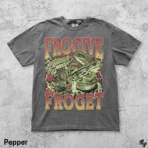 Pu&ograve; includere: T-shirt color pepe con una grafica di diverse rane verdi e la scritta "FROGive FROGet" in un font retr&ograve;. La maglietta ha un aspetto vintage e usurato.