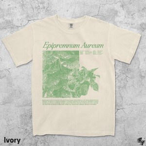 May include: Ivory-colored t-shirt with a green botanical illustration and the text "Epipremnum Aureum." The design features a detailed drawing of a plant with leaves and stems. The shirt also includes additional text describing the plant's origin.