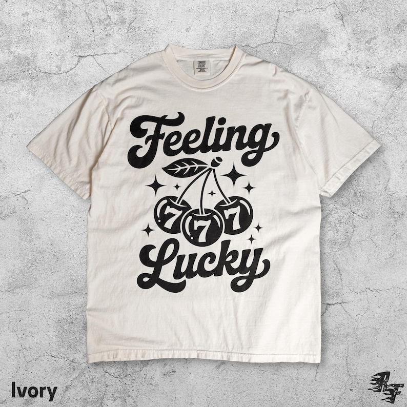 May include: Ivory-colored t-shirt with the words "Feeling Lucky" in a bold, black script. The design features a cluster of cherries with the number "7" inside each cherry. The shirt is a casual, unisex style.