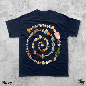 Puede incluir: Camiseta azul marino con un dise&ntilde;o en espiral hecho de varias piedras, conchas y un helecho coloridos. La palabra "Navy" est&aacute; impresa en la esquina inferior izquierda.